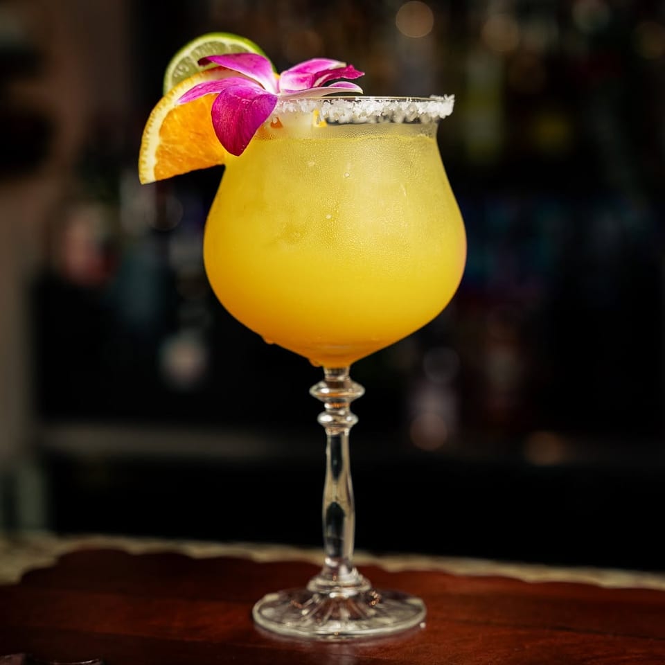 Vibrant yellow cocktail with citrus slices and a pink orchid on a salt-rimmed glass.