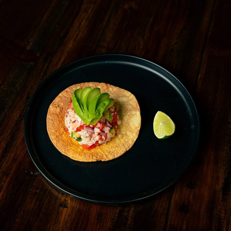 A tostada topped with ceviche and sliced avocado, served with a lime wedge on a black plate.