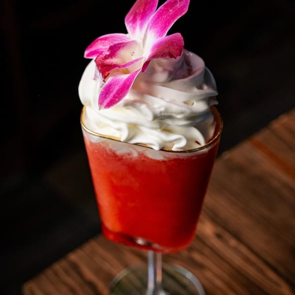 Vibrant red frozen cocktail topped with whipped cream and a pink orchid on a wooden table.