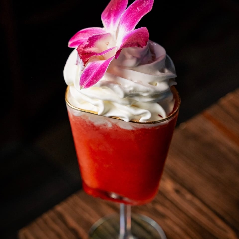 Vibrant red frozen cocktail topped with whipped cream and a pink orchid on a wooden table.