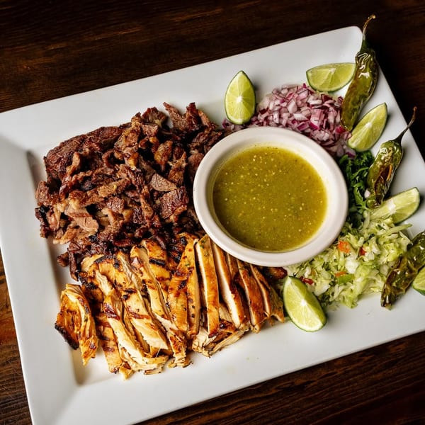 Platter with grilled chicken, steak, green salsa, limes, onions, peppers, and cabbage slaw on a wooden table.