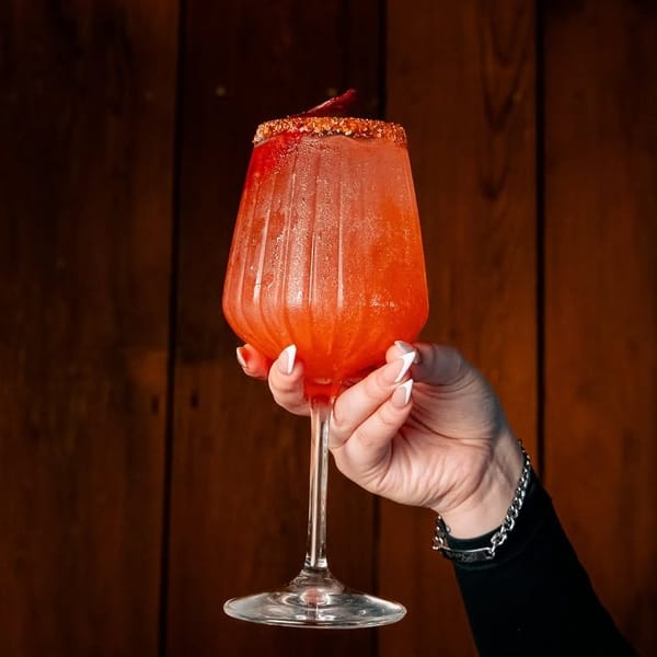 A hand holds a vibrant red cocktail in a textured glass, rimmed with spices, against a wooden background.