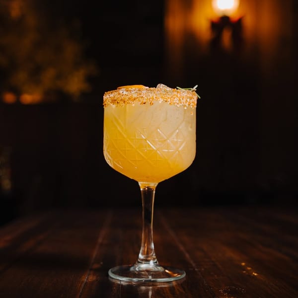 Yellow cocktail in a patterned glass with a salted rim and garnish on a dark wooden table.