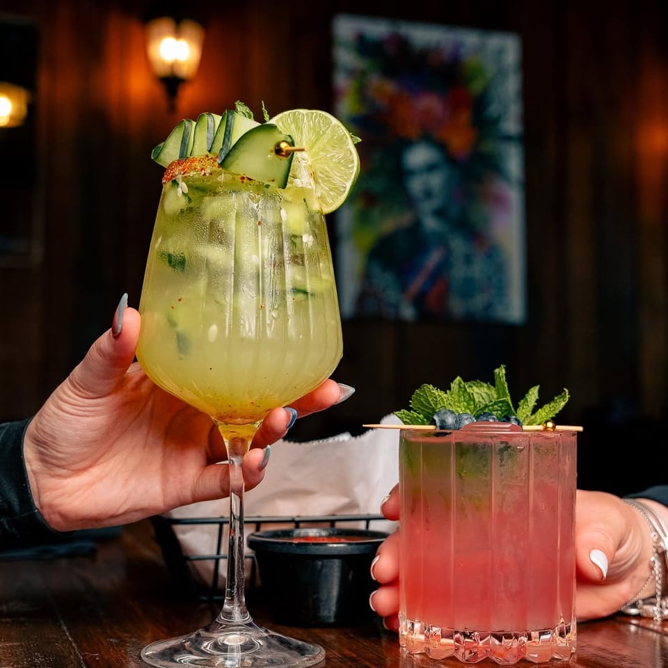 Two hands holding cocktails, one cucumber-lime and one berry-mint, on a dark wooden table.