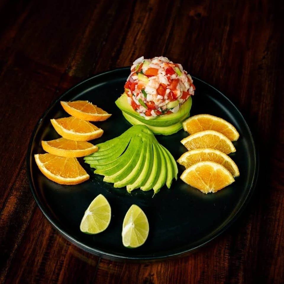Ceviche in an avocado bowl, surrounded by sliced avocado, orange wedges, and lime on a black plate.